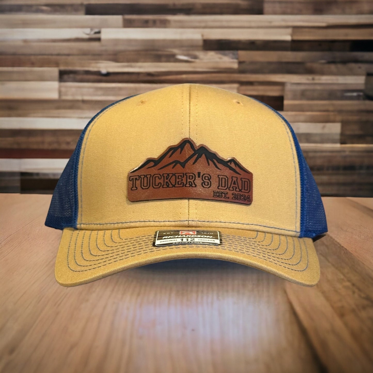 May include: A yellow and blue trucker hat with a brown leather patch that says 'Tucker's Dad Est. 2024' and a mountain range design.
