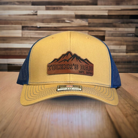 May include: A yellow and blue trucker hat with a brown leather patch that says 'Tucker's Dad Est. 2024' and a mountain range design.